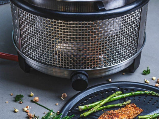 Cobb gassgrill i buk med stekeplate liggende på bordet ved siden av grillen med ferdiggrillet asparges og et kjøttstykke