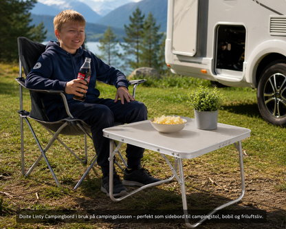 Gutt sitter ved Doite Linty bord med en cola utenfor en bobil ved en vakker innsjø, perfekt sidebord for camping