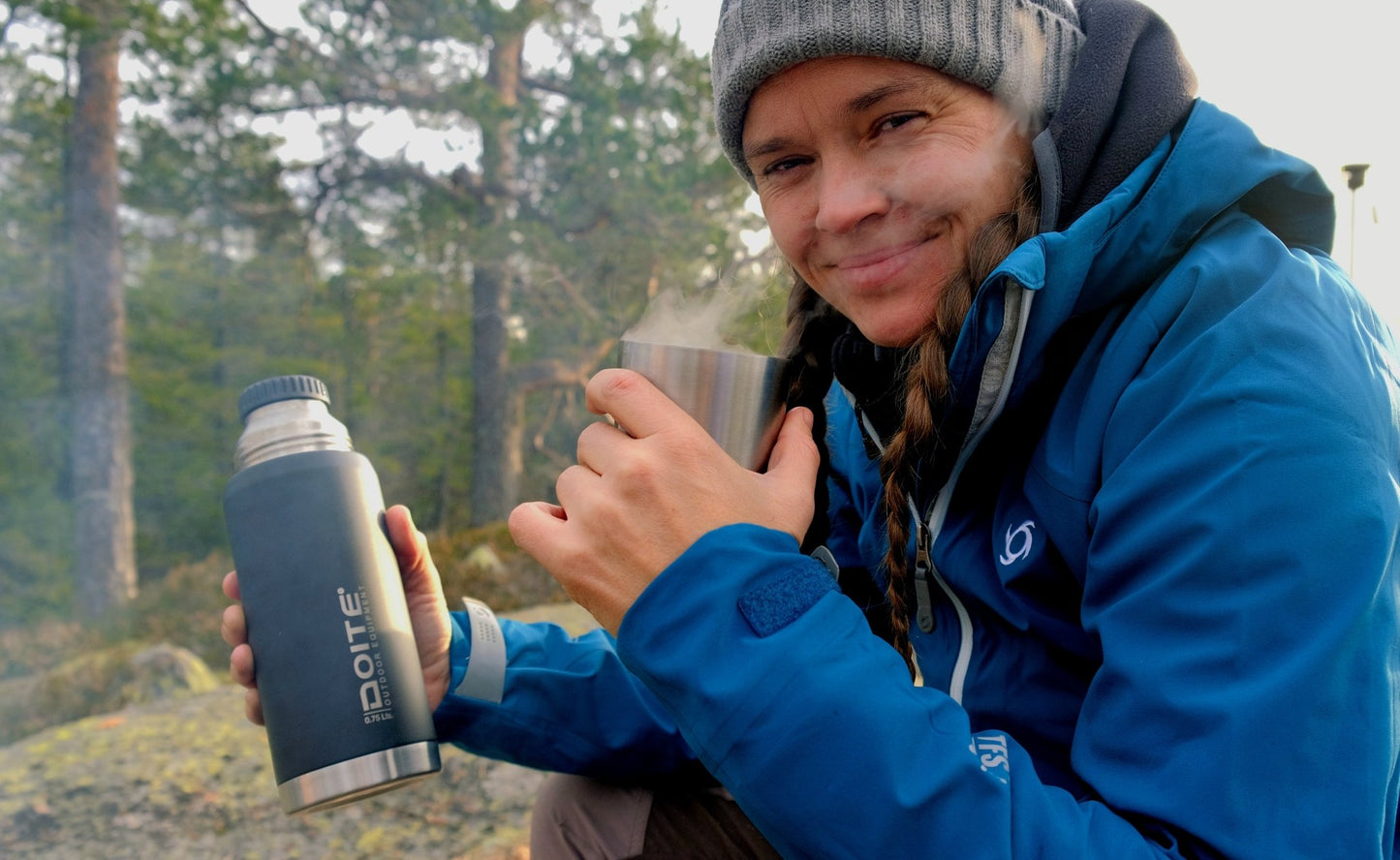 nærbilde av en kvinne som holder en doite termos i høyrehånd og drikker av termoskoppen med venstre, ikledd en blå jakke fra Doite, i et skogsområde